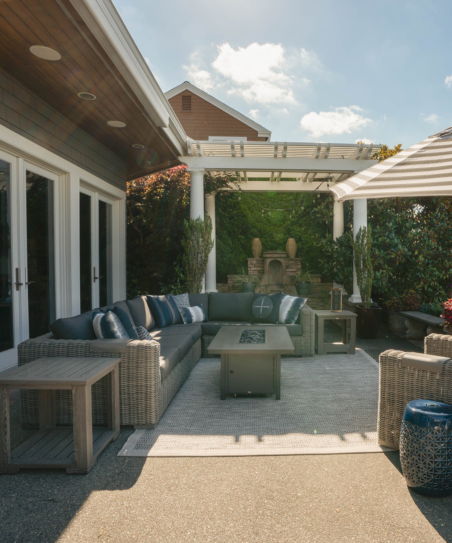 A large woven outdoor sectional with dark grey cushions flanked by two aluminum side tables and a fire pit table in the center