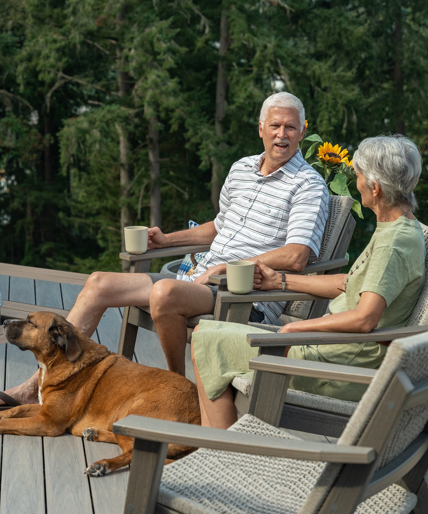 Two people sitting in patio chairs on a deck drinking coffee while a dog lays at their feet.